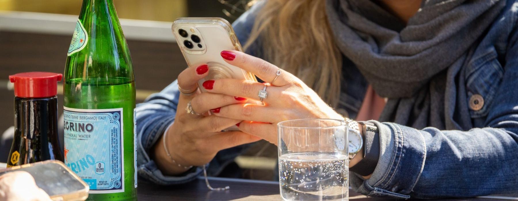 a woman holding a phone with a bottle and a glass on the table she's sitting at